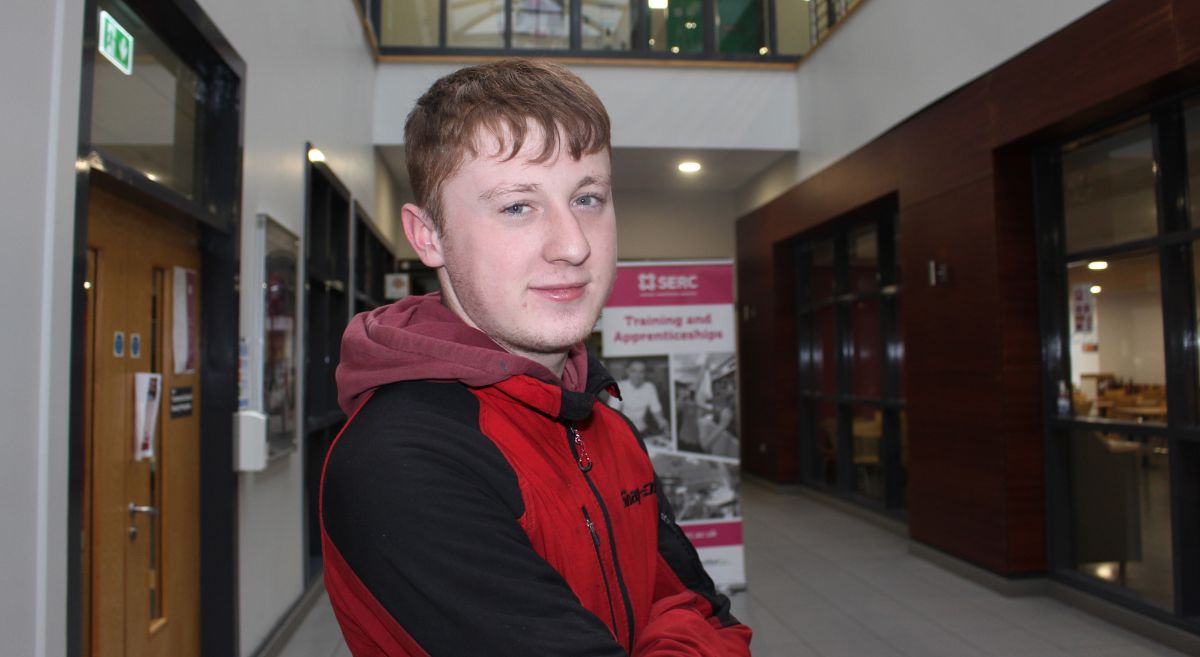 male wearing black and red hoodie smiling to camera, pictured in college corridor